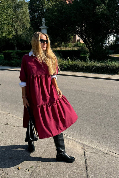 Kvinde i bordeaux kjole med T-shirt ærmer, sorte støvler, solbriller og taske.