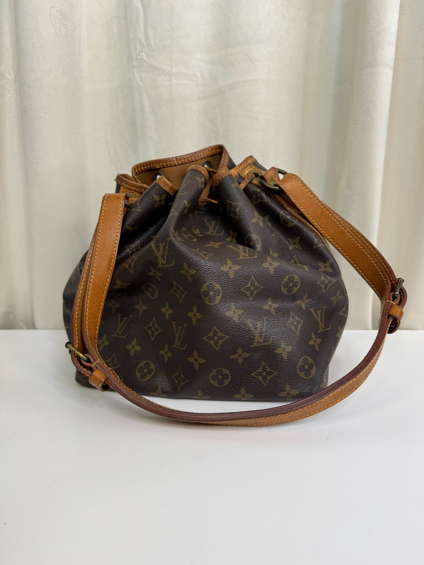 Louis Vuitton skuldertaske med monogram, brun læderrem og toplukning på lys baggrund.