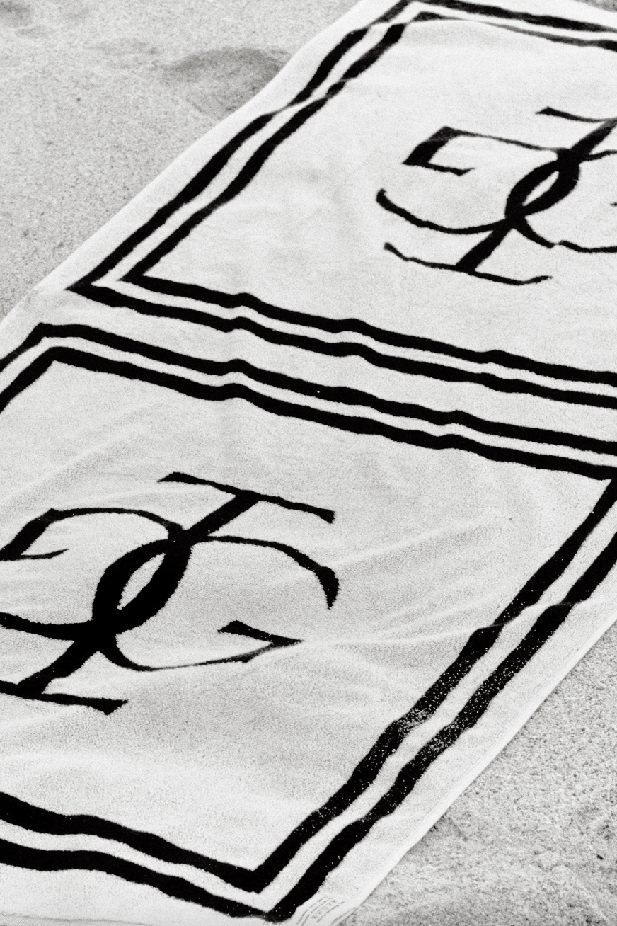 Håndklæde med sort-hvidt design, logo i midten, striber langs kanten, placeret på sand.